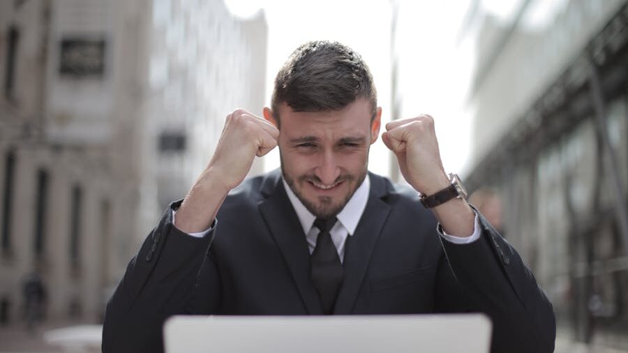 A man in a suit sitting outdoors in front of a laptop, raising his fists with a tense expression, symbolizing emotional intensity or frustration at the end of a digital interaction.