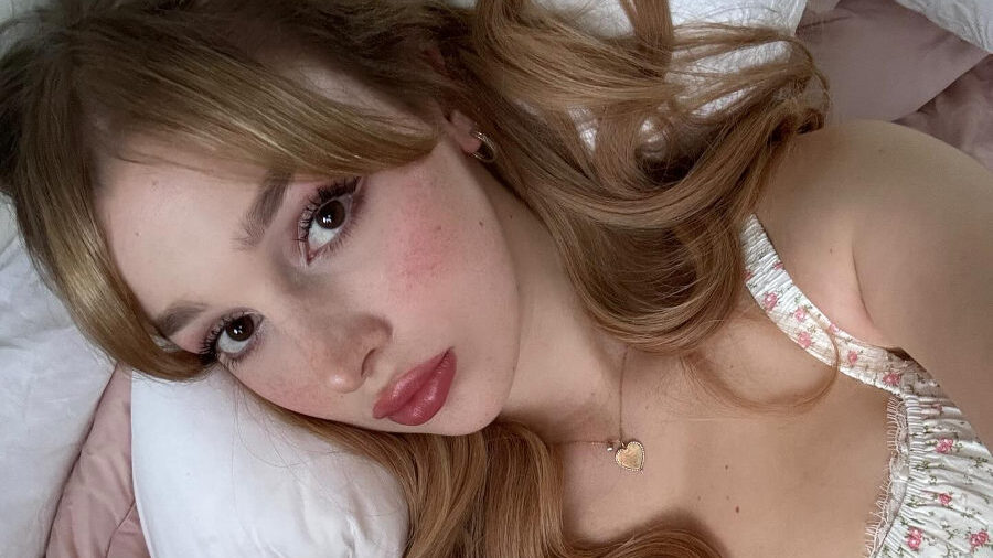 Grace Hyland lies on a bed, taking a selfie with her head resting on a white pillow. She wears a floral-printed dress and a heart-shaped necklace. Her makeup is soft and romantic, and her long hair flows over her shoulders. The photo is inviting and softly lit, with a caption promising a surprise in the next slide.