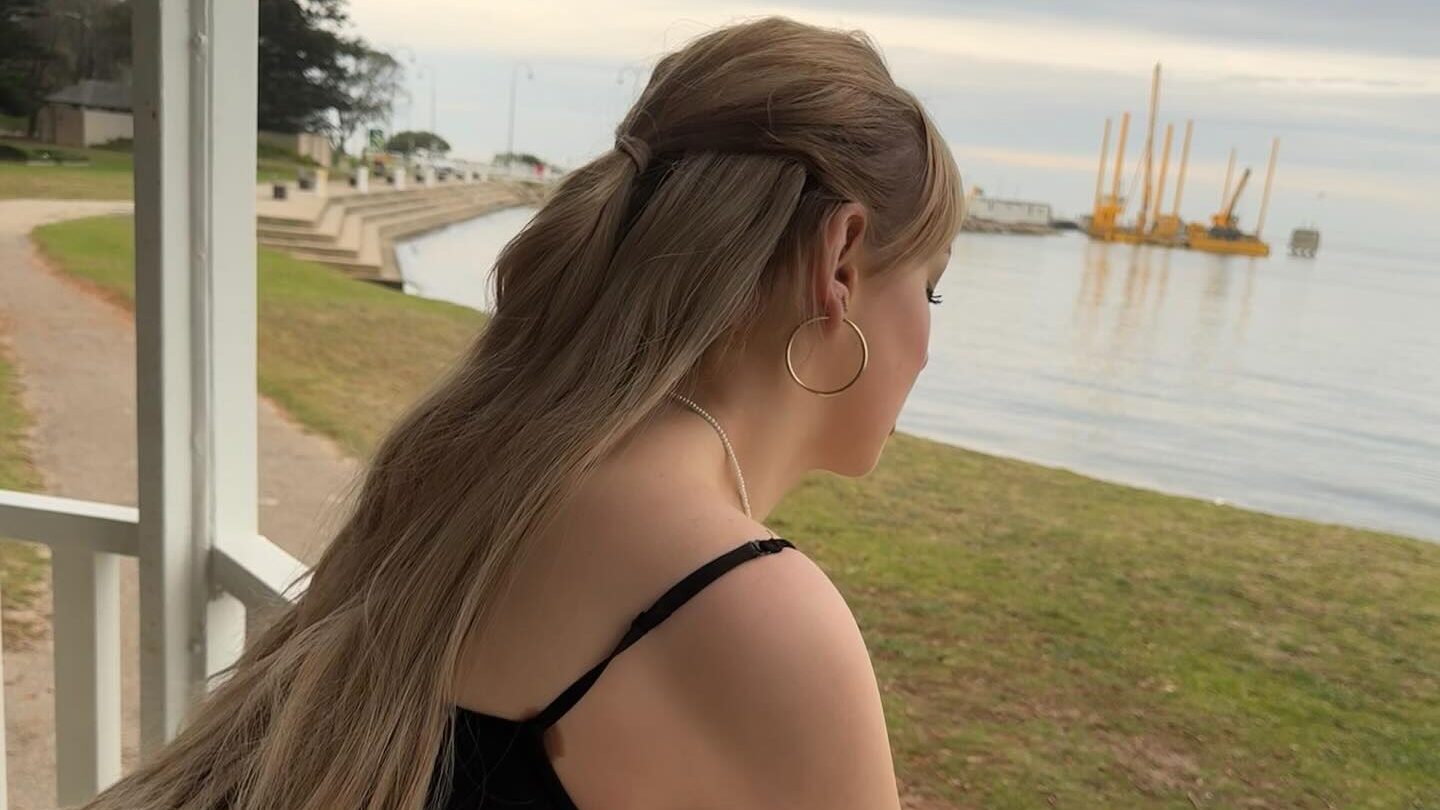 Grace Hyland stands on a white wooden balcony, leaning forward as she looks peacefully toward the water. Her long, blond hair is half-tied back, and she wears a black top with thin straps and large hoop earrings. The scene is tranquil, with calm water, overcast skies, and a few yellow structures in the distance, evoking a mood of introspection and serenity.