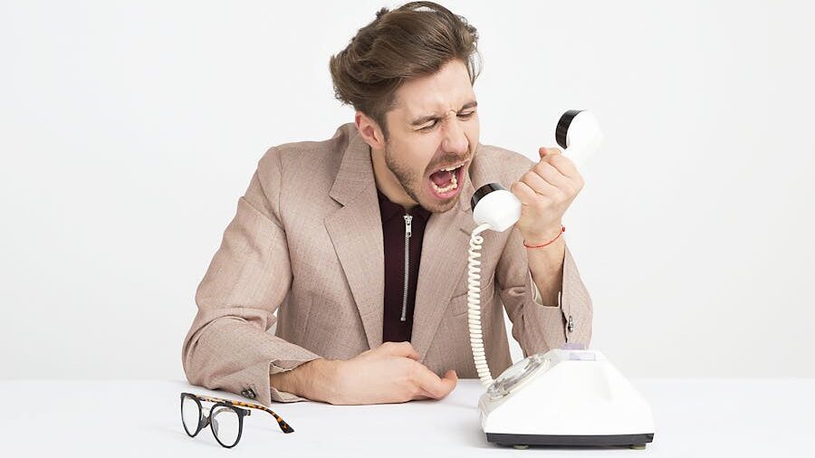 A man in a beige blazer angrily yells into a vintage white telephone while sitting at a table, showing visible frustration and impatience; his eyeglasses rest beside him.
