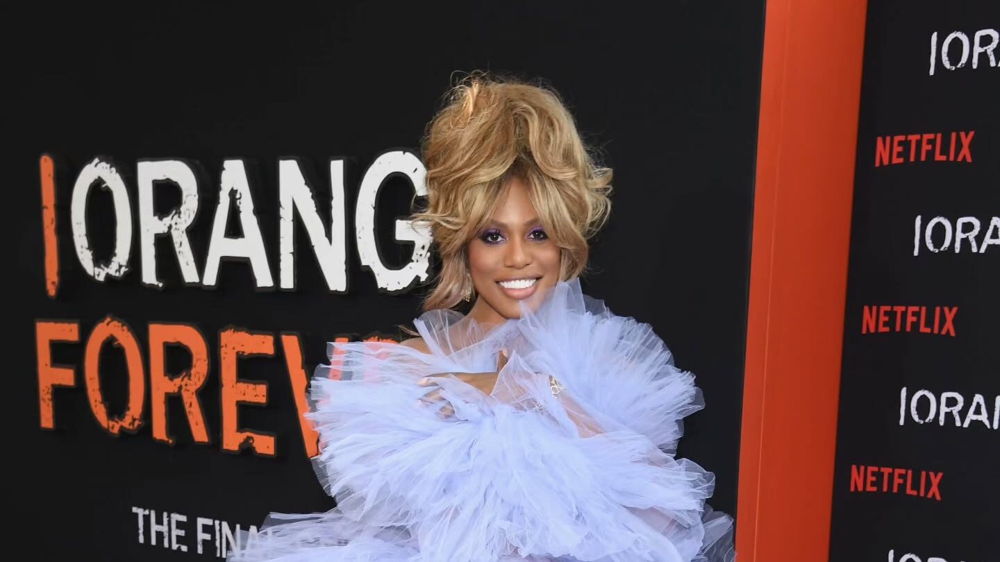 Laverne Cox poses in a voluminous pale lavender ruffled gown on the red carpet for the Orange Is the New Black anniversary celebration. She smiles brightly in front of a black and orange Netflix backdrop that reads “ORANGE FOREVER.”