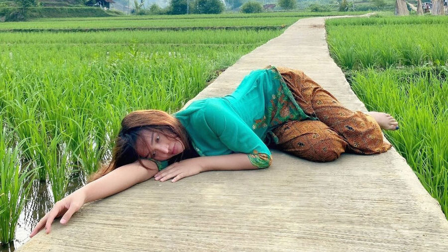 Lucinta Luna lies in a relaxed pose on a narrow concrete path surrounded by green rice paddies in Subang, West Java. She wears a bright green kebaya and patterned sarong, with tropical mountains and scattered trees in the distance.