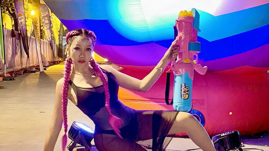 Lucinta Luna kneels on a street at the S2O Songkran Festival in Bangkok, holding a large water gun in front of a giant rainbow-colored inflatable backdrop. She wears a sheer black mesh outfit with braided pink hair and chunky black shoes.