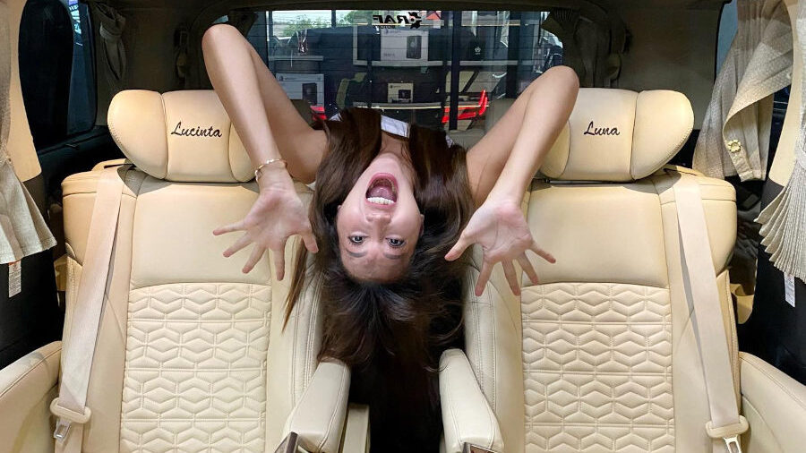 Lucinta Luna hangs upside down between two cream-colored luxury seats inside her customized Alphard van. Her name is embroidered on the headrests, and she’s making a playful expression with her hands spread out.