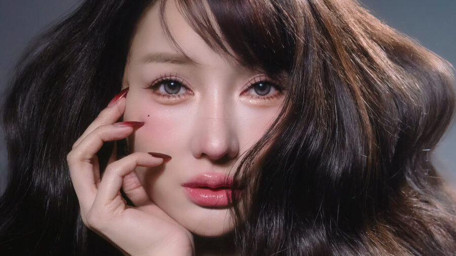 Close-up portrait of Lucinta Luna with voluminous, softly waved brown hair cascading over one shoulder. Her hand is gently placed on her cheek, with manicured nails and a serene expression.