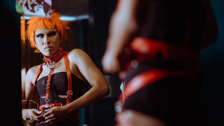 Person with vivid orange hair in a black and red harness outfit looks into a mirror with intensity, questioning gaze and strong makeup under dramatic lighting.