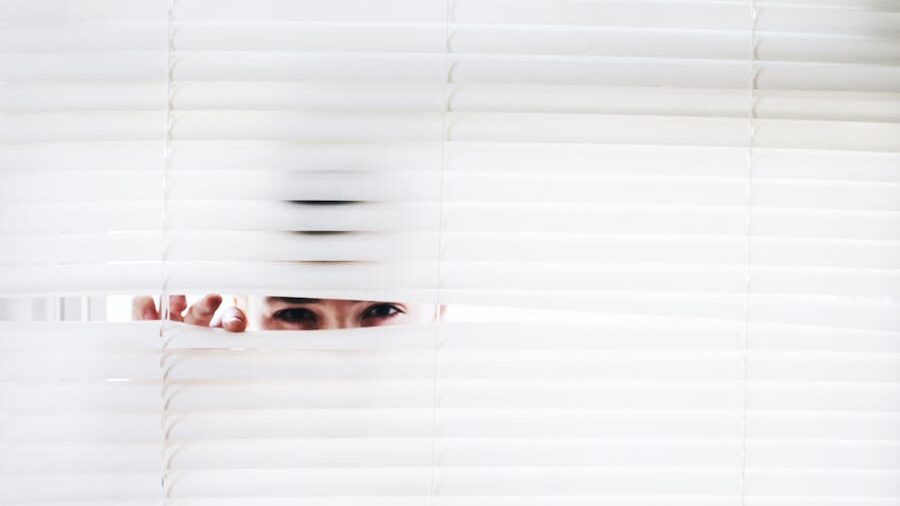 A person peeks through slightly opened white blinds, revealing only their eyes and part of their hand, suggesting secrecy or surveillance.