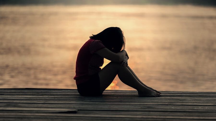 A person sits alone on a wooden dock at sunset, knees pulled to their chest, in a reflective or emotional posture symbolizing solitude and the emotional weight of past relationships.