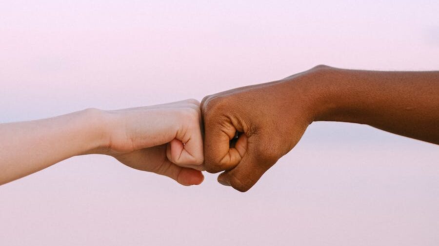 Close-up of two fists, one light-skinned and one dark-skinned, gently bumping each other against a soft pink background, symbolizing unity and understanding.