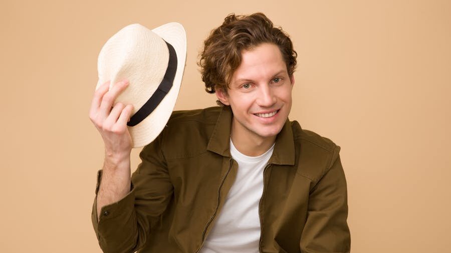 Smiling man in a white shirt and green jacket, tipping his straw hat politely in front of a neutral background.
