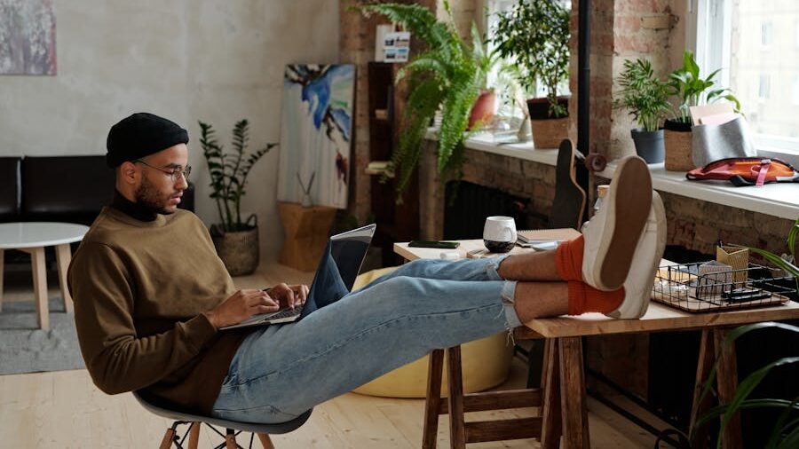 A man sits casually at a desk with his feet up, working on a laptop in a stylish, plant-filled room—representing laid-back modernity and the repetition of aesthetic norms.