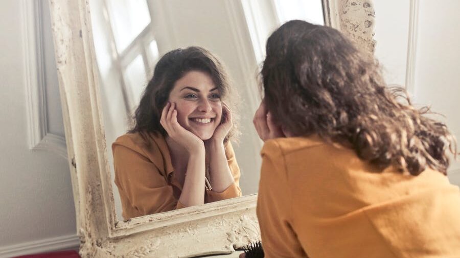 A smiling woman looking at herself in the mirror, resting her face in her hands, expressing self-love and joy.