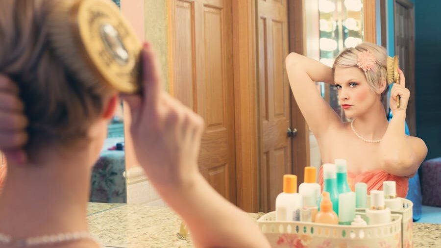 A woman with a limb difference is brushing her hair in front of a well-lit mirror, surrounded by beauty products, with confidence and poise.