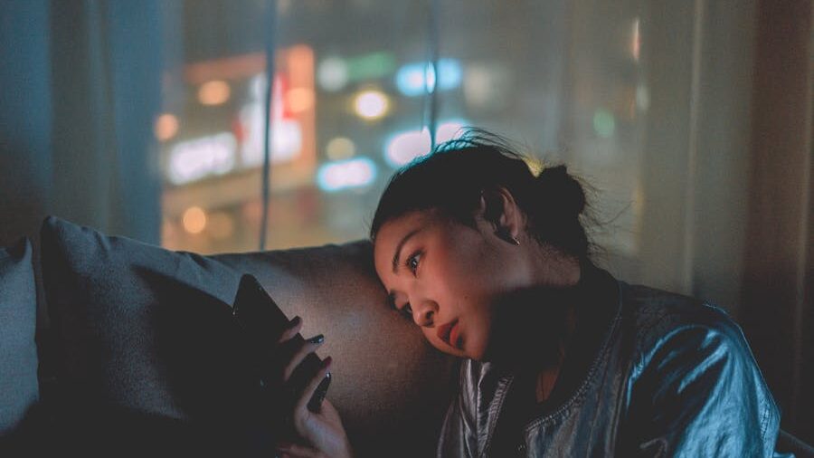 A woman sitting alone in a dimly lit room at night, leaning her head on a pillow and looking at her phone with a pensive expression.
