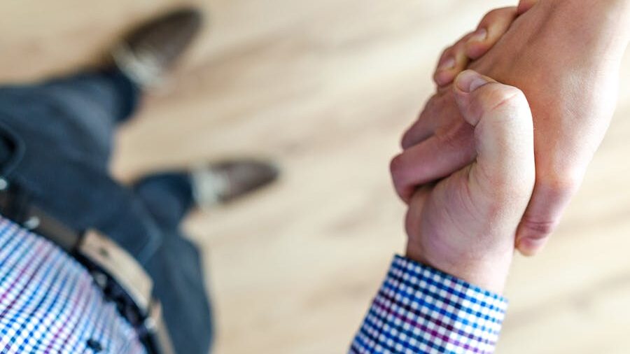 Close-up of two people shaking hands, one in a checkered shirt, viewed from above, standing on a wooden floor.