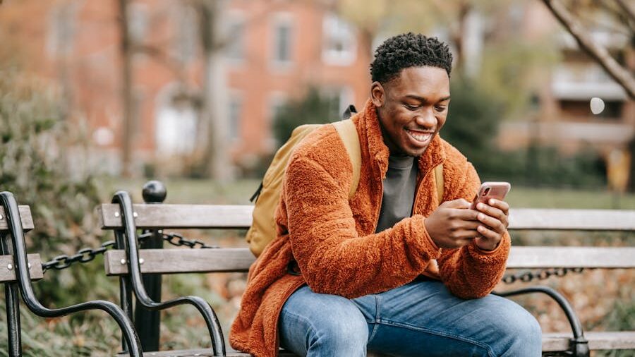 A young man sitting on a park bench in autumn, smiling as he looks at his phone, wearing a warm orange jacket and a backpack.