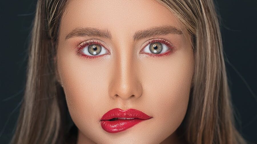 Face of a woman with striking grey eyes, red lip gloss, and flawless skin against a dark background.