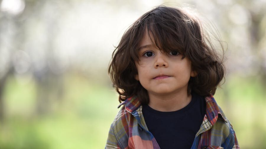 Child with serious expression and tousled hair, standing in a blurred green orchard setting.