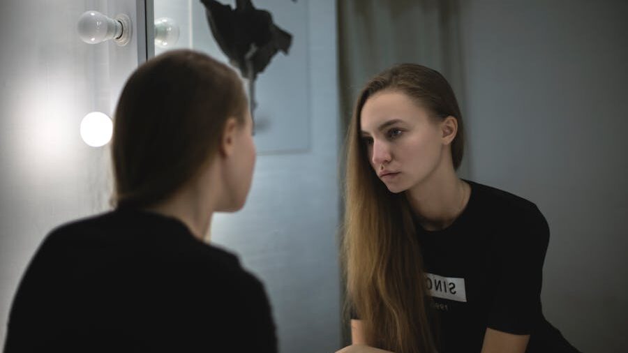 Pensive woman in a black shirt sitting in front of a lit mirror, lost in introspective thoughts.
