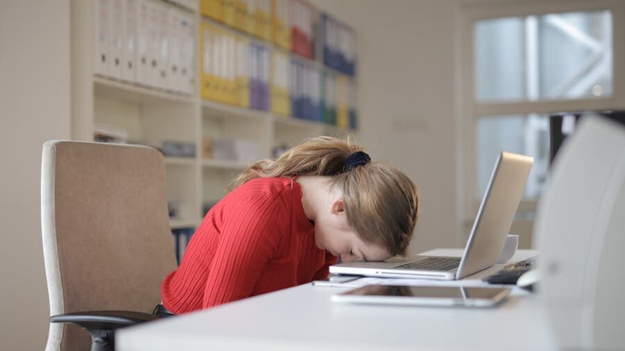 Tired person resting their head on a laptop at a white desk, surrounded by tech and office folders.