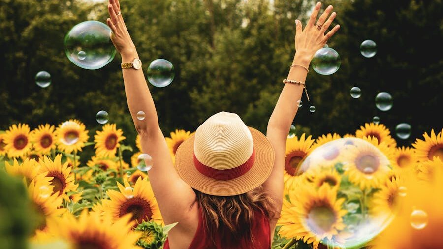 A woman in a straw hat stands with arms raised in a sunflower field, surrounded by soap bubbles under a clear summer sky.