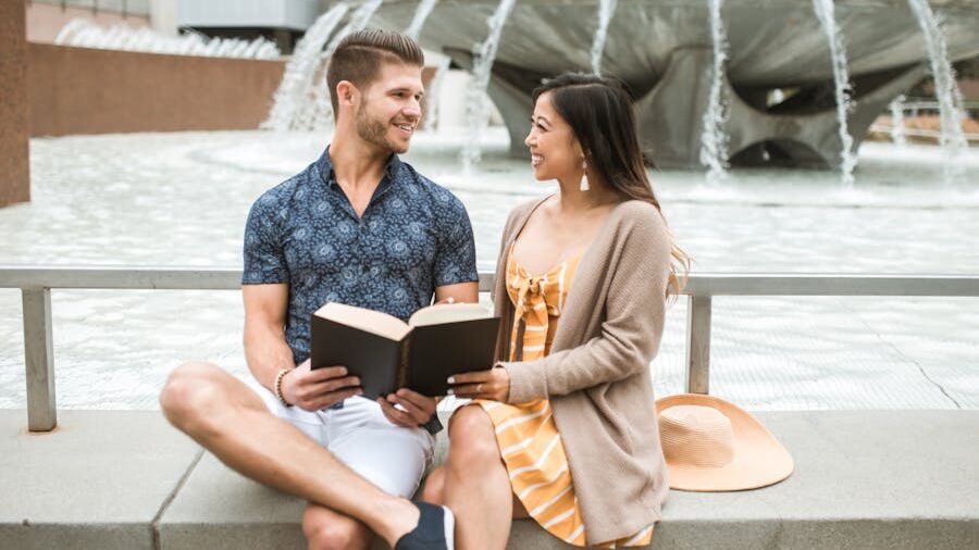A smiling couple sits by a city fountain sharing a book, enjoying a warm moment of connection and calm outdoors.