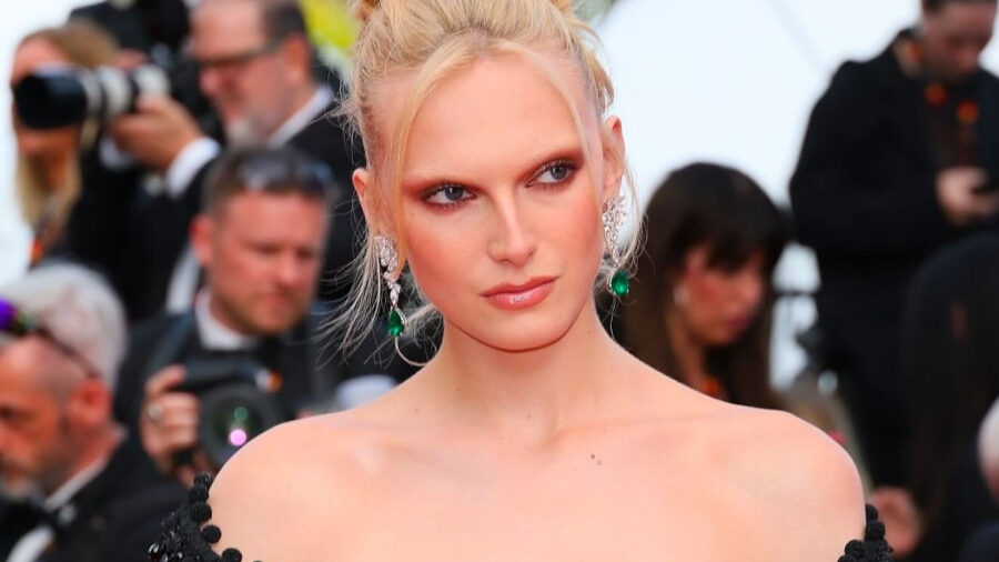 Alex Consani on a red carpet wearing a black off-the-shoulder gown adorned with beaded embellishments, with an elegant updo and bold green earrings.