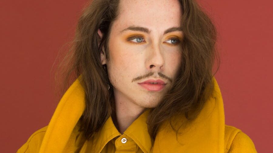 A person with long brown hair, orange eyeshadow, and a thin mustache poses confidently in a yellow shirt against a red background.