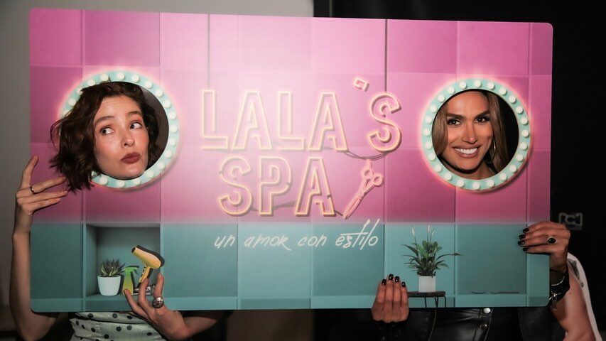 Isabella Santiago and co-star through cutouts of a colorful spa promo board.