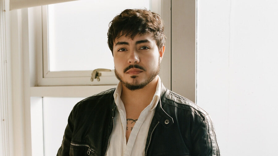 Ryan Jacobs Flores standing indoors in front of a window in Los Angeles, wearing a leather jacket and white shirt.