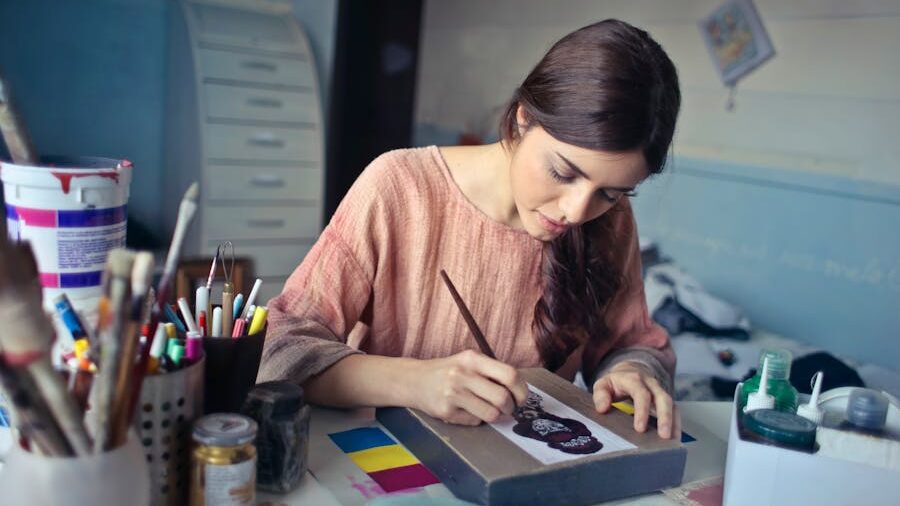Artist working intently on a painting in a room filled with pens, brushes, and paint jars.