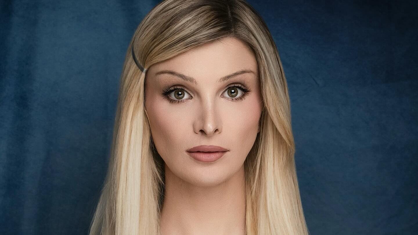 Dylan Mulvaney photographed for a glam yearbook-style portrait, lit softly in a studio with a dark blue backdrop.