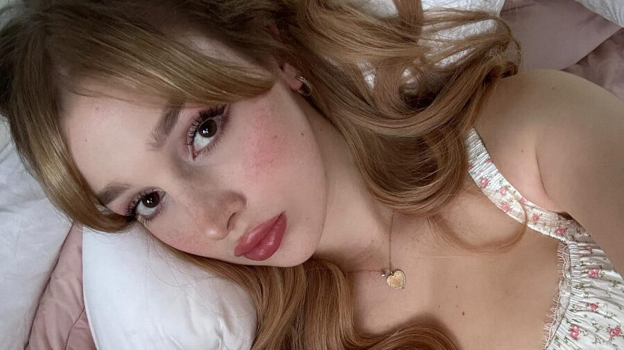 Grace Hyland lies on a bed, taking a selfie with her head resting on a white pillow. She wears a floral-printed dress and a heart-shaped necklace. Her makeup is soft and romantic, and her long hair flows over her shoulders. The photo is inviting and softly lit, with a caption promising a surprise in the next slide."