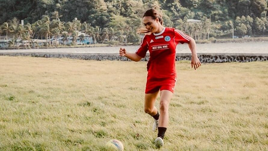 Jaiyah Saelua in red uniform kicking a soccer ball on grass field at Fagaalu Park, surrounded by palm trees.