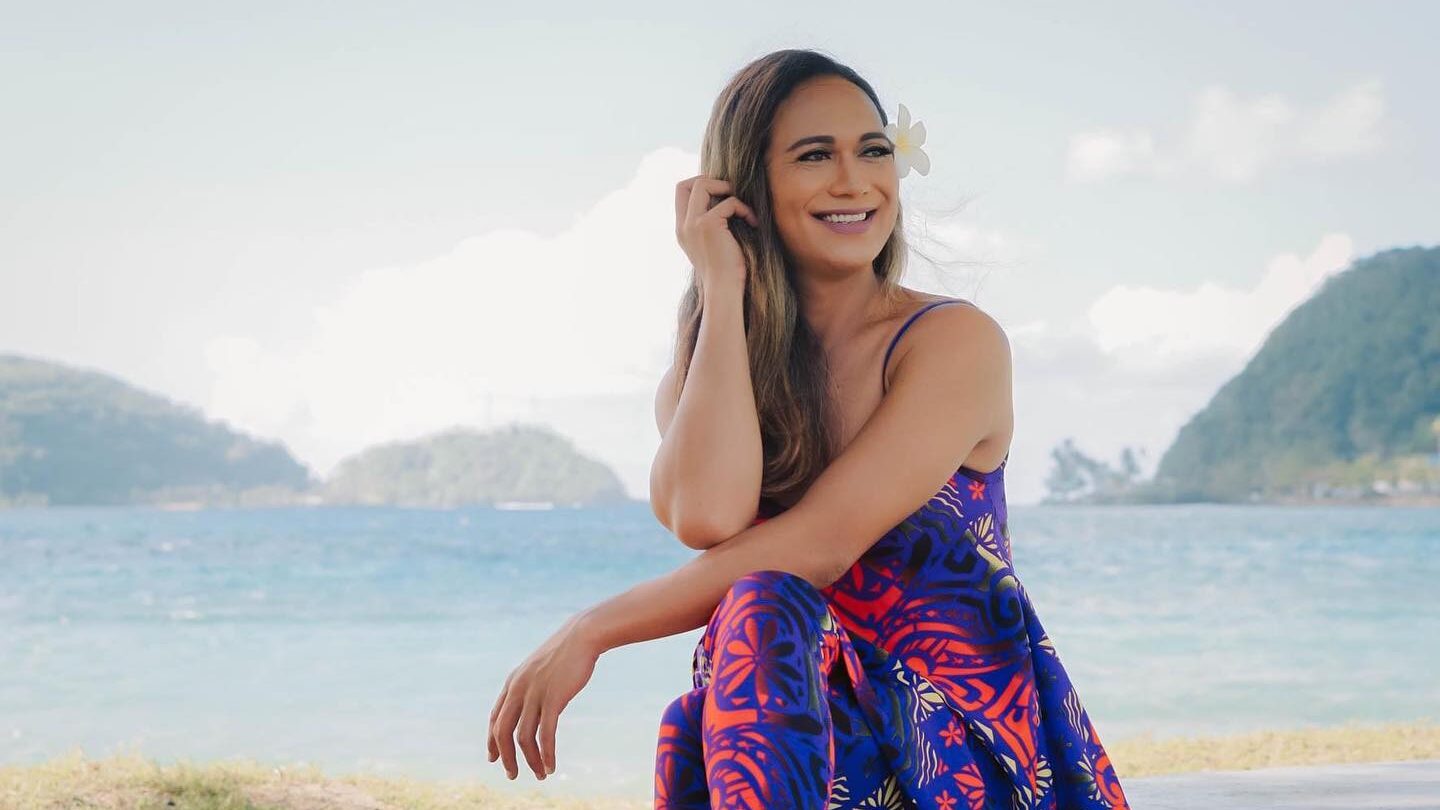 Jaiyah Saelua sitting on grass by the ocean in Samoa, wearing a colorful dress.
