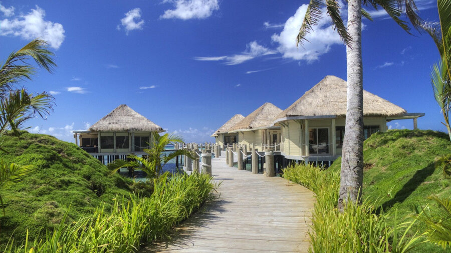 Seaside resort with palm trees and stilted bungalows under a bright blue sky and scattered clouds.