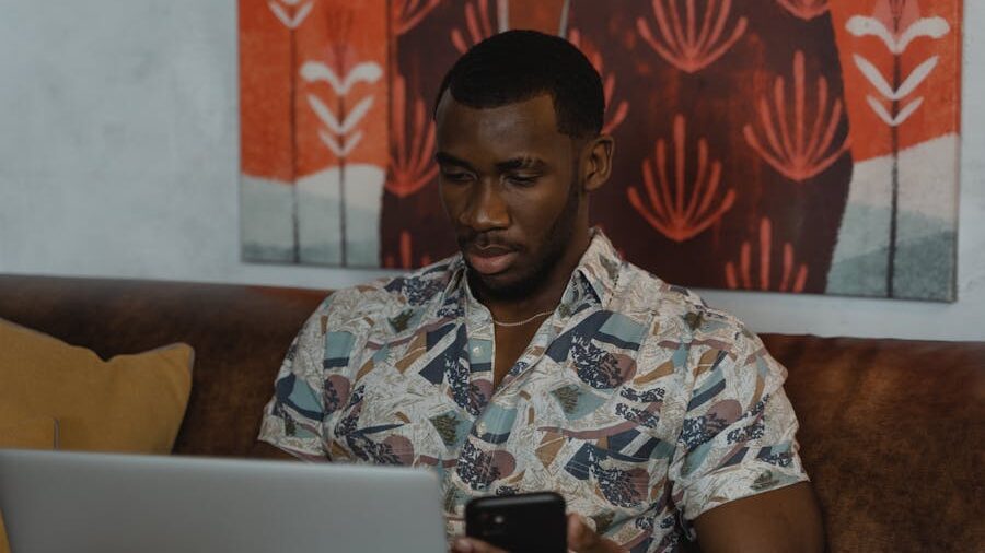 A man sitting on a couch looking at his phone with a laptop in front of him, in a warm and modern interior setting.