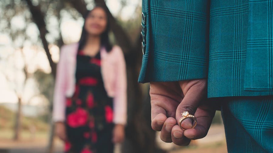 A man hides a ring behind his back, facing a woman in the background, suggesting an upcoming marriage proposal.