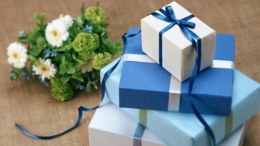 Three blue and white gift boxes stacked with satin ribbons beside a bouquet of green and white flowers on a burlap surface.