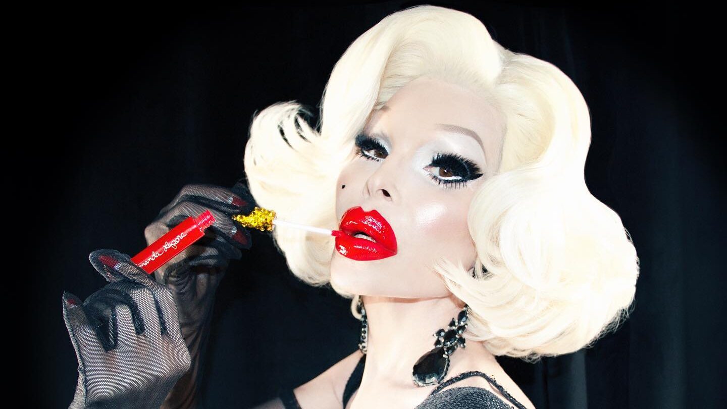 Amanda Lepore with platinum blonde hair and red lip gloss, holding the gloss applicator in front of a black background.