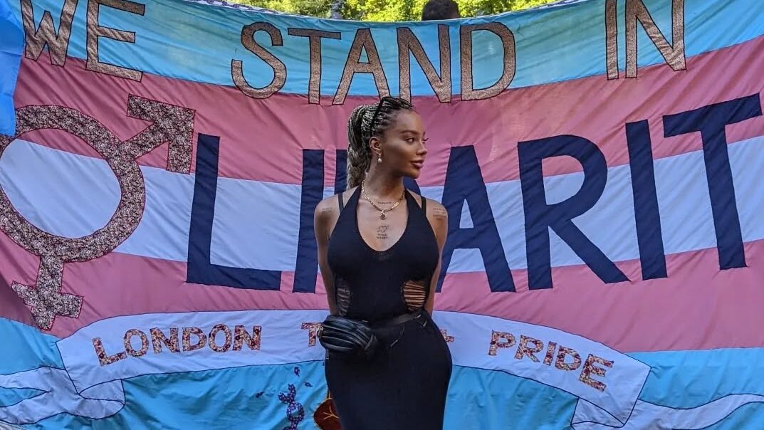 Munroe Bergdorf in a long black dress posing in front of a trans flag banner reading “We Stand in Solidarity” at Pride 2022.