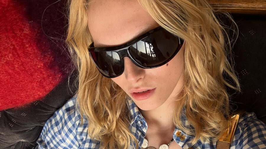 Alex Consani sitting in a relaxed pose, wearing a blue gingham shirt, white mini skirt, dark sunglasses, and shoulder bag.