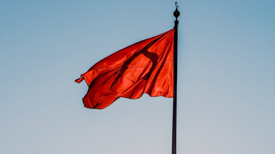 A red flag mounted on a tall black pole, blowing in the wind against a cloudless blue sky.