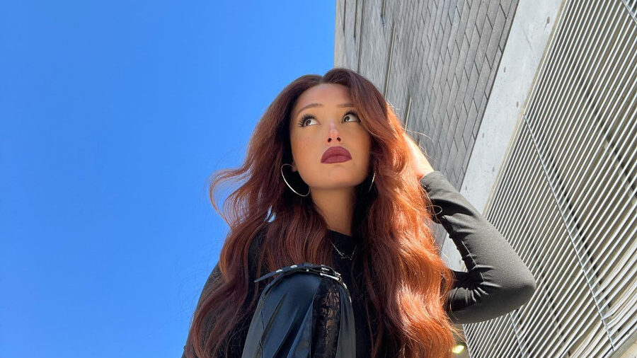 Zoey Luna poses outdoors in black leather pants and long red waves, sitting on a crate under sunlight.