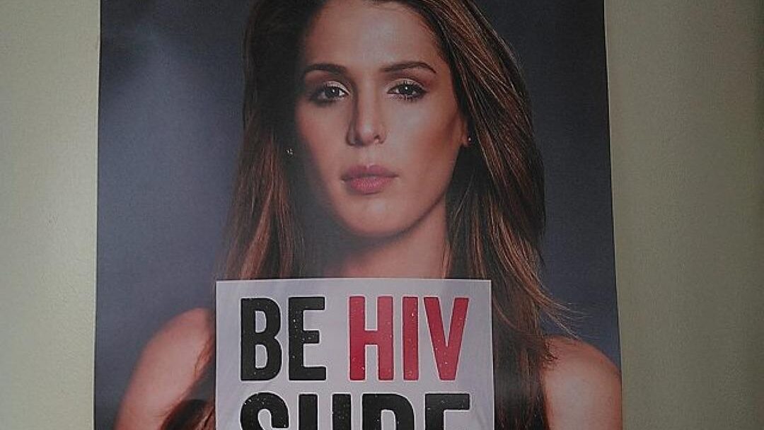 Carmen Carrera posing in an HIV awareness poster holding a sign reading “Be HIV Sure,”.