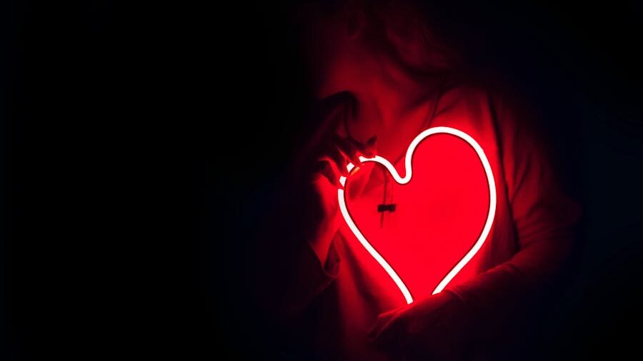 Red neon heart lighting up the chest of someone in a dark background.