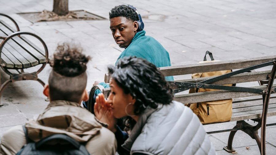 Three people sitting on benches; two whisper while one looks back with suspicion.