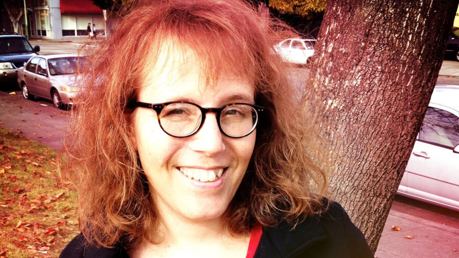 Julia Serano smiling outdoors near a tree, wearing glasses and a black coat with a red top, parked cars behind her.