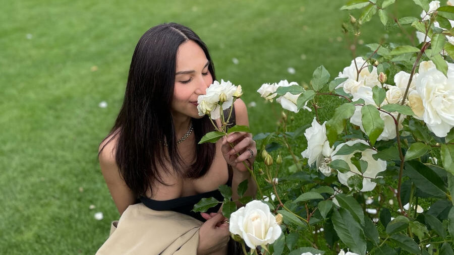 Dena Rachman kneels beside blooming white roses in York Museum Gardens with historic stone ruins in the background.