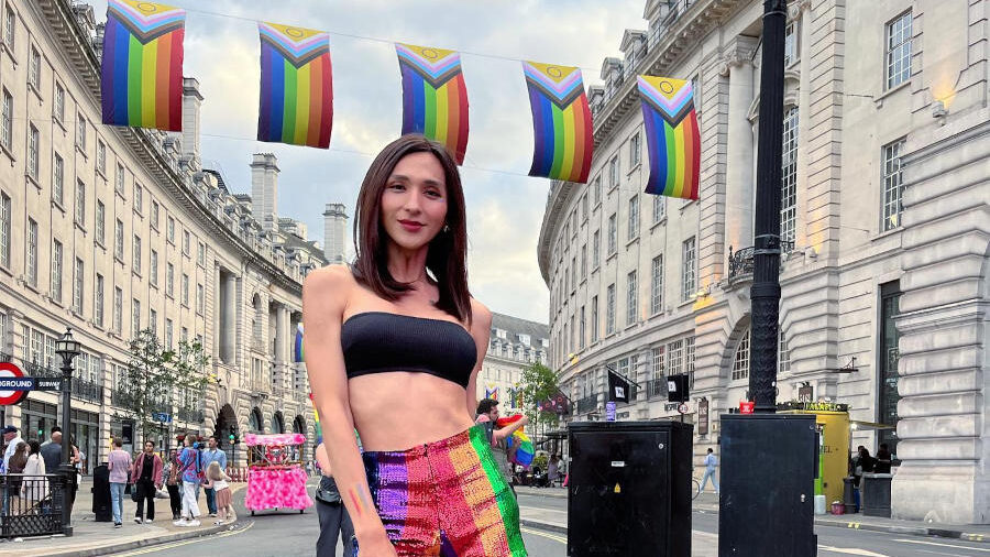 Dena Rachman poses on a London street during Pride, wearing rainbow shorts beneath colorful pride flags.